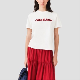 Model wearing Maje white crew-neck tee with red C&ocirc;te d'Azur slogan, styled with a red tiered midi skirt and blue bag.