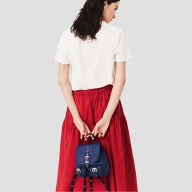 Model carrying Maje metallic mini backpack with buckle detail, styled with white tee and red midi skirt