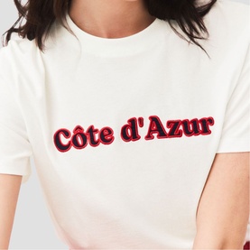 Model wearing Maje white crew-neck T-shirt with red and black C&ocirc;te d'Azur slogan print on chest.