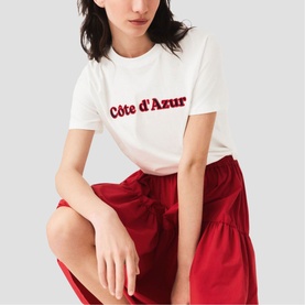 Model wearing Maje white C&ocirc;te d'Azur slogan T-shirt with red metallic text, styled with a red wide-leg skirt.