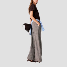 Model carrying Maje dark brown disc crossbody bag, styled with grey wide-leg trousers, brown tee and light blue shirt tied at waist
