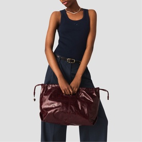 Model carrying Maje large burgundy patent leather tote bag with drawstring top and gold-tone hardware, styled with navy tank and wide-leg trousers
