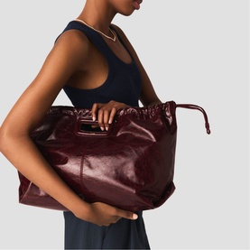 Model carrying Maje bag in deep burgundy patent leather, large slouchy silhouette with drawstring top closure, held under arm