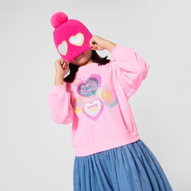 Model wearing Billy Blush pink heart-print sweatshirt with sequin detail, styled with blue tulle skirt and pink pom-pom beanie hat