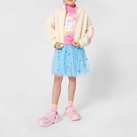 Model wearing Billy Blush blue star-print tulle skirt, styled with cream jacket, pink graphic top and pink Crocs