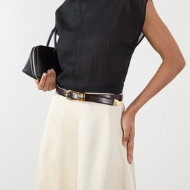 Model with Dehanche Hollyhock Leather Belt in deep brown, gold-tone rounded buckle, worn over cream skirt; model carrying a black clutch bag.