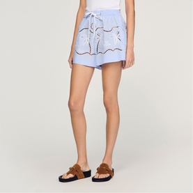 Model wearing Sandro sky blue broderie anglaise shorts with drawstring waist, styled with tan strappy flat sandals.