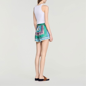 Model wearing Sandro green and purple printed mini skirt with white sleeveless top and tan flat sandals, rear view