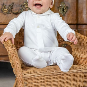 Baby wearing Emile Et Rose white all-in-one with footed trousers, bow detail, and matching hat, seated in wicker chair