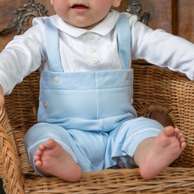 Baby wearing Emile Et Rose pale blue dungaree outfit with white long-sleeve bodysuit, seated in wicker basket