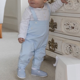 Baby wearing Emile Et Rose pale blue dungarees with white long-sleeve top and blue boots, standing by a white dresser.