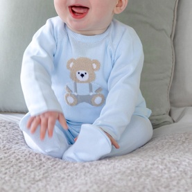 Baby wearing Emile Et Rose pale blue all-in-one with brown teddy bear appliqu&eacute; detail, seated on a sofa cushion