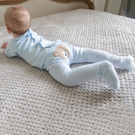 Baby wearing Emile Et Rose pale blue all-in-one sleepsuit with teddy bear appliqu&eacute; detail on back, lying on chunky knit blanket.