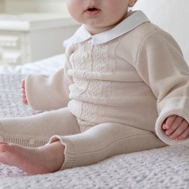 Model wearing Emile Et Rose stone knit all-in-one babygrow with white collar detail, seated on white bedding