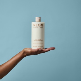 Neom Organics Body Wash in large cream bottle with rose gold logo, held aloft on a single outstretched hand against blue background