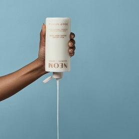 Neom Organics Body Wash in cream pump bottle held upside down dispensing product against light blue background