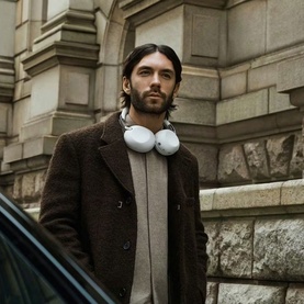 Model wearing white over-ear headphones around neck, styled with a brown wool coat and grey knit, urban stone building backdrop