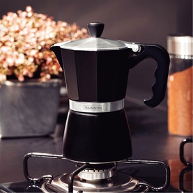 Tramontina black stovetop moka pot coffee maker on a gas hob, with silver band and black handle, plants in background.