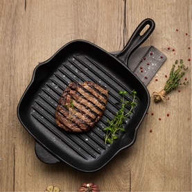 Tramontina black cast iron grill pan with ridged surface, styled with a grilled steak and fresh herbs on a wooden surface.