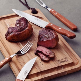 Tramontina brown wooden-handled cutlery set displayed on wooden board with sliced steak, including steak knives, fork, and carving fork.