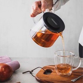 Tramontina clear glass teapot with black lid pouring tea into a glass mug on a wooden board, with an apple in the background