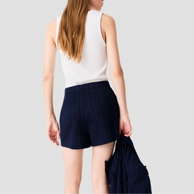 Model wearing Maje navy textured shorts with white sleeveless top, rear view, holding a navy jacket