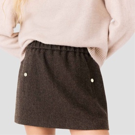 Model wearing Maje dark brown mini skirt with elasticated waist and pearl button details, styled with a blush pink knit sweater.