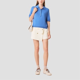 Model wearing Maje blue knit polo top with cream double-breasted mini shorts, styled with beige bag and trainers