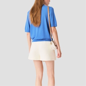 Model carrying Maje bag in multi, worn crossbody, styled with blue knit top and cream shorts, rear view