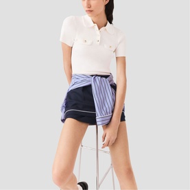 Model wearing Maje white cropped polo shirt and navy shorts with blue striped shirt tied at waist