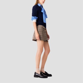 Model wearing Maje multi plaid mini skirt styled with navy knit, blue shirt with bow tie, and black loafers