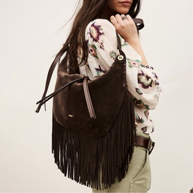 Model carrying Bash brown suede fringe shoulder bag with long tassel trim, worn over floral print blouse with khaki trousers