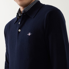 Model wearing Gant Shield Logo Fine-Knit Polo Sweater in navy, three-button placket, chest logo, styled with checked shirt collar visible beneath.