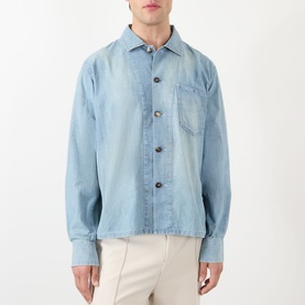 Model wearing JW Anderson Slant Pocket Denim Shirt in pale blue with washed effect, pointed collar and button fastenings, styled with cream trousers.