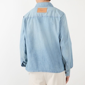 Model wearing JW Anderson Slant Pocket Denim Shirt in pale blue, rear view showing washed denim, tan logo patch, styled with cream trousers.