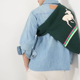 Model wearing JW Anderson Slant Pocket Denim Shirt in pale blue, styled with a green crossbody bag featuring equestrian motif.