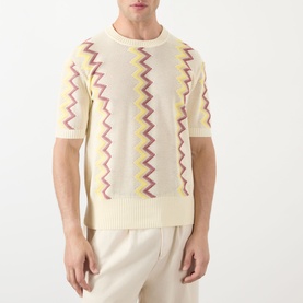 Model wearing JW Anderson Chevron Pattern Sweater in ivory with purple and yellow zigzag detail, crew neck, short sleeves, paired with cream trousers.