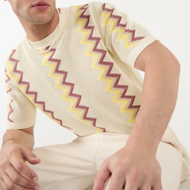Model wearing JW Anderson Chevron Pattern Sweater in ivory with multicolour zigzag knit, crew neck, short sleeves, styled with cream trousers.