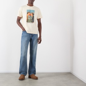 Model wearing JW Anderson Mid-Rise Straight Leg Jeans in mid blue faded wash, styled with a cream graphic tee and tan shoes.
