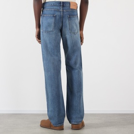 Model wearing JW Anderson Mid-Rise Straight Leg Jeans in mid blue faded wash, rear view showing leather logo patch and five-pocket styling.