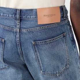 Model wearing JW Anderson Mid-Rise Straight Leg Jeans in mid blue, close-up of rear showing leather logo patch and back pocket detail.