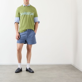 Model wearing JW Anderson Hiking Chino Shorts in steel blue with belt loops, styled with green logo tee, blue shirt and black loafers.
