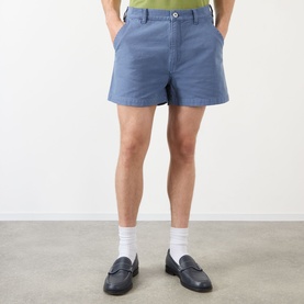 Model wearing JW Anderson Hiking Chino Shorts in steel blue cotton with belt loops and button fastening, styled with white socks and black loafers.