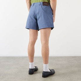 Model wearing JW Anderson Hiking Chino Shorts in steel blue with back logo patch, styled with white socks and black loafers.