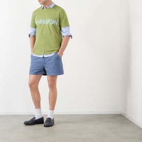 Model wearing JW Anderson Hiking Chino Shorts in steel blue with belt loops, styled with green logo tee, blue shirt and black loafers.