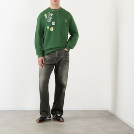 Model wearing JW Anderson Scattered Logo Sweatshirt in green with crew neck and letter graphics, styled with dark grey jeans.