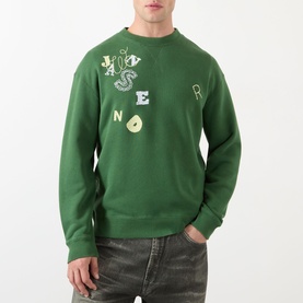 Model wearing JW Anderson Scattered Logo Sweatshirt in green with crew neck, ribbed trims, and scattered letter graphics. Styled with grey jeans.