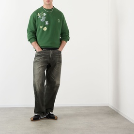 Model wearing JW Anderson Scattered Logo Sweatshirt in green with crew neck, styled with dark wash jeans and black shoes.