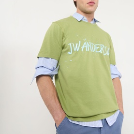 Model wearing JW Anderson Circular Logo T-Shirt in leaf green with blue graphic logo print, styled over a blue striped shirt.