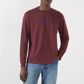 Model wearing JW Anderson burgundy long-sleeve cotton T-shirt with blue logo embroidery at chest, styled with light blue jeans.
